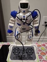 Front view of Ruko 6088 programmable robot with wired remote control placed in front, showing detailed buttons on remote and robot design.