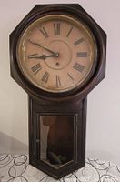Wall mounted grandfather style clock with octagonal wooden frame, Roman numeral face, two clock hands, and lower glass window for pendulum.