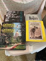 Four coffee table books placed on fabric, including two copies of Georgia Land of Many Dreams, two Beatles books, and National Geographic Society anniversary book.