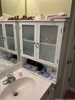 Photo of white bathroom wall cabinet mounted above sink with frosted glass doors and lower open shelf, showing context and surrounding sink area