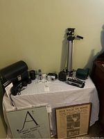 Wide shot showing multiple cameras, lenses, a tripod, and camera bag on a white cloth-covered table, including a sign with text and a folder.