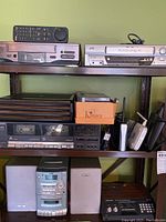 Two JVC VHS players on upper shelf, one with remote, plus Sony cassette player with box of cassettes, RCA audio system with speakers and cassette/CD player on lower shelf.