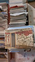 Photo showing multiple stacks of postcards with a close-up on a postcard depicting an ancient fresco wall art with horses and human figures.