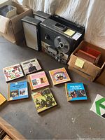 Vintage reel-to-reel tape recorder on the floor with two matching speakers and multiple stereo tape albums scattered around, some in cardboard boxes.
