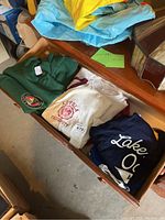 Four sweatshirts including green with circular emblem, white with Loyola University text, navy blue with partial text, and royal blue with Lakeshore Oaks text.