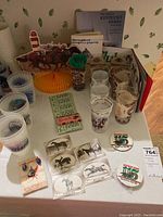 Wide view of the collection of Kentucky Derby glassware, plastic cups, tickets, pins, brochures, and horse figurines on a table against floral wallpaper.