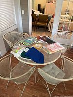 White round wrought iron mesh table with four matching wrought iron mesh chairs around it. Chairs have light gray padded cushions. Various fabric items placed on table.