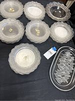 Eight frosted glass Lalique Honfleur plates in two stacks of four next to a larger frosted glass bowl and oval crystal platter on black cloth background.