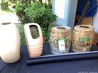 Display of two ceramic barrels, two large vases, and rolled black wallpaper on a black cloth outdoor table