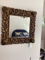 Full view of square mirror with ornate carved wooden frame hanging on wall, showing floral and scroll patterns.