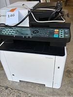 Front-right angled view of Kyocera multifunction printer with control panel, power cord, and USB cable coiled on top.