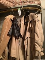 Three women's coats hanging on a bar: tan shearling jacket, black coat, beige trench coat.