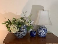 Full view of all three items on wooden surface: blue and white floral bowl with artificial flowers, small blue and white ginger jar, and ginger jar lamp with white shade.