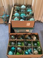 Two cardboard storage boxes stacked on a plastic bin showing assorted blue glass ornaments and a star topper leaning against the wall.
