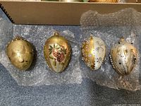 Four silver and gold egg-shaped and round ornaments laid on bubble wrap showing detailed hand-painted and glitter accents