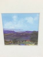 Close-up photo of watercolor artwork showing purple mountains, blue sky, clouds, some birds flying, and small buildings with trees by a water body.