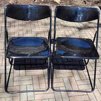 Front view of two black folding chairs with metal frames and glossy plastic seats and backs, showing both chairs side by side.
