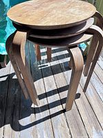 Three stacked vintage wooden stools outdoors on weathered wooden floor, showing round seats and curved bentwood legs