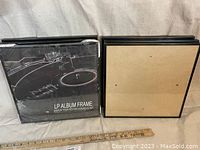 Stack of six black square LP album frames, one wrapped in shrink-wrap packaging with product image and text on front.