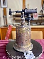 Front and side views of antique brass torch light showing brass cylindrical tank, cast iron valve and burner mechanism, red wooden handle, and heavy patina and wear indicative of age.