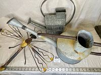 Three items: large metal watering tin with rust spots, smaller metal watering can, and steel wire dragonfly garden decor with yellow accents, visible on a cream background with a measuring ruler in front.