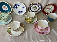 Wide view showing seven assorted bone china tea cups and matching saucers with floral designs and gold trim
