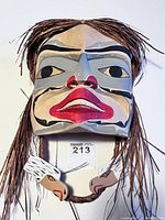 Front view of carved and painted female portrait mask showing facial details, signature, and attached hair-like woven strands and bird head chin strap.