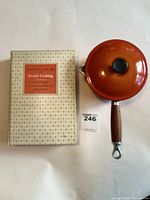 Photo of vintage Le Creuset orange sauce pan with lid and the vintage book next to it on white background.