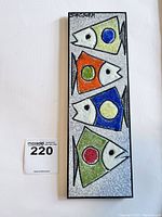 Rectangular ceramic wall plaque with four colorful stylized fish arranged vertically. Artist signature visible in top left corner. Good lighting shows surface texture and colors.