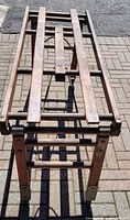Top down view of the collapsible wooden stand laid out flat on a brick driveway, showing wood slats and folding mechanism.