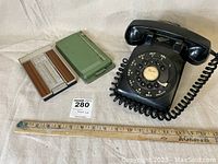 Black rotary dial phone with coiled handset and two vintage Rolodex card holders, one chrome with wood grain panels and one green plastic case.
