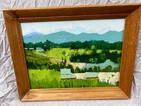 Framed oil-on-canvas painting showing an expansive lush valley with farm buildings and distant mountains under a cloudy sky.