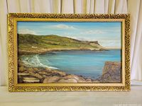 Full view of the framed oil painting depicting a coastal scene with rocks, green hills and blue sea under a partly cloudy sky.