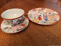 Photo of a Nippon hand-painted teacup and saucer alongside a hand-painted decorative plate with traditional scene and floral patterns on a wooden table.