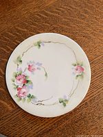 Top view of round ceramic trivet showing hand painted pink roses, green leaves, and gold accent lines on white ceramic.