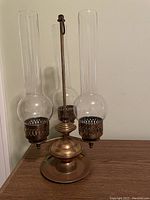 Photo showing a brass lamp with three clear glass chimneys, perforated brass holders, and a circular base on a neutral background.