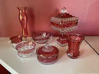 Seven cranberry glass dishes and vases arranged on a white surface against a pink wall background. Items include varying shapes and styles with a rich red cranberry hue and clear textured glass detail.