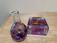 Small hand blown glass vase with purple mottled pattern and a rectangular decorative glass box with purple sides and multicolored segmented lid on wooden table.