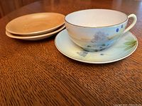 Hand painted Nippon tea cup with matching saucer and two beige plates from occupied Japan on wooden table.