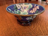 Close-up photo of Japanese cloisonné enamel bowl with floral and leaf patterns in blue background, on wooden carved stand.