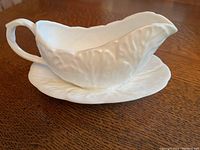 White English bone china Wedgwood pitcher with embossed leaf pattern sitting on matching saucer on wooden surface.