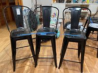 Photo showing three black metal barstools with curved backs and square seats arranged side by side on a wooden floor.
