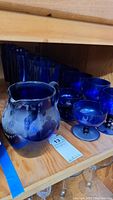 Photo of large blue glass pitcher and goblet-style glasses arranged on wooden shelf.