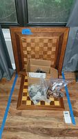 Wooden chess board with a checkerboard pattern in varying wood shades, a cardboard box, and plastic bags containing chess pieces.