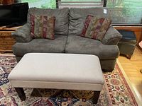 Gray fabric couch with two multicolored patterned cushions seen from front side, sitting on floral patterned rug with wooden furniture around.
