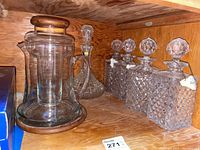 Six decanters arranged on a wooden shelf showing the glass pitcher with wood base and four square diamond cut decanters with faceted stoppers.
