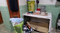 Overview photo showing dog crate topped with white cloth, surrounded by two green laundry baskets filled with blankets and towels, metal bowls on top, and a metal trashcan in background.