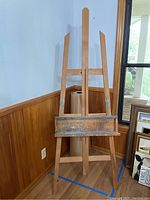 Side view of tall wooden easel with adjustable shelf. Easel shows signs of use with paint marks on the shelf bracket. Large roll of brown kraft paper standing behind the easel.