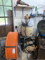 Photo shows large orange industrial fan with visible wear, sitting on the floor near a green metal rolling cart with items stored on it, and a Milwaukee grinder/sander placed on a wooden shelf above the cart.