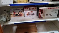 Shelf view of Apple Peeler Corer Slicer in box, Zoku Quick Pop Maker box, and KitchenAid Gourmet Pasta Press box with six shaping discs.
